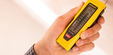 Hand holding a yellow Stanley moisture meter against a white surface, displaying a reading of 26.