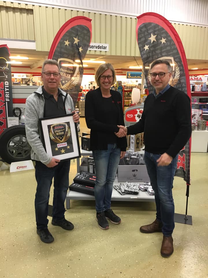 Three people indoors, one holding framed award, two shaking hands by display. Red and black banners in background.