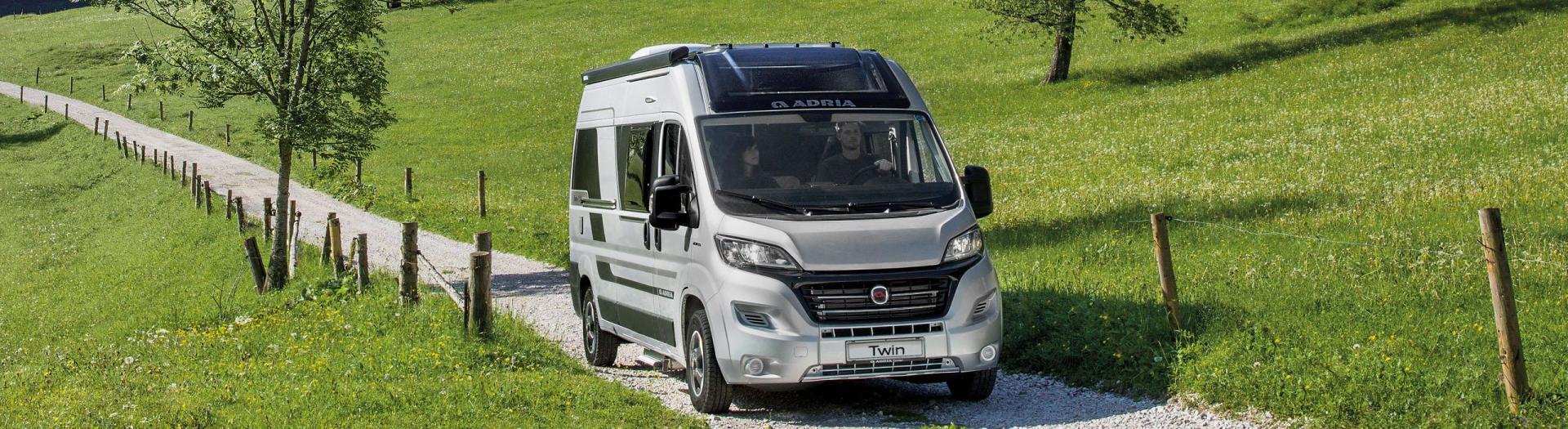 A silver RV driving on a gravel road through a grassy field.
