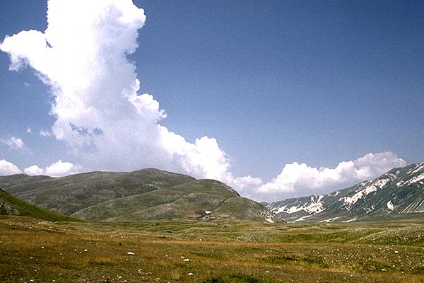 Ein weites, grasbewachsenes Tal mit Bergen unter einem strahlend blauen Himmel, übersät mit großen, weißen Wolken.