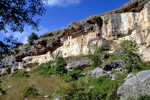 Felsige Klippen mit in den Stein gehauenen Gebäuden, grünem Laub und einem klaren blauen Himmel.