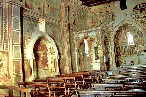Innenraum einer alten Kirche mit verblassten Fresken an den Wänden und gewölbten Türen. Holzbänke säumen den Boden.