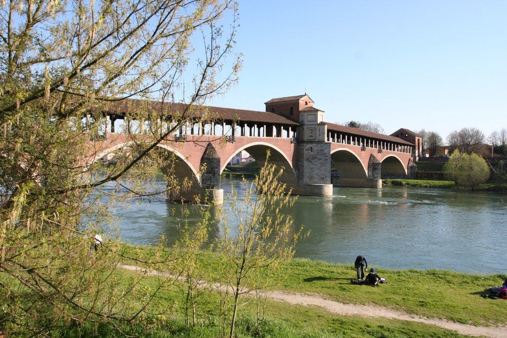 Eine überdachte Brücke mit roten Bögen überspannt einen Fluss in Pavia, Italien. Menschen entspannen am grasbewachsenen Ufer.