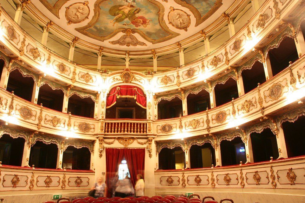 Kunstvoller, mehrstufiger Theaterinnenraum mit goldenen Akzenten, einer Bühne mit rotem Vorhang und einer bemalten Decke. 