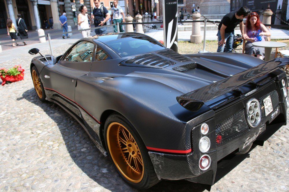 Dunkelgrauer Supersportwagen Pagani Zonda mit goldenen Rädern, der im Freien geparkt ist; in der Nähe sind Menschen zu sehen.
