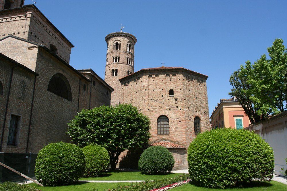Ein Steingebäude mit einem runden Backsteinturm und einem Garten davor; blauer Himmel.
