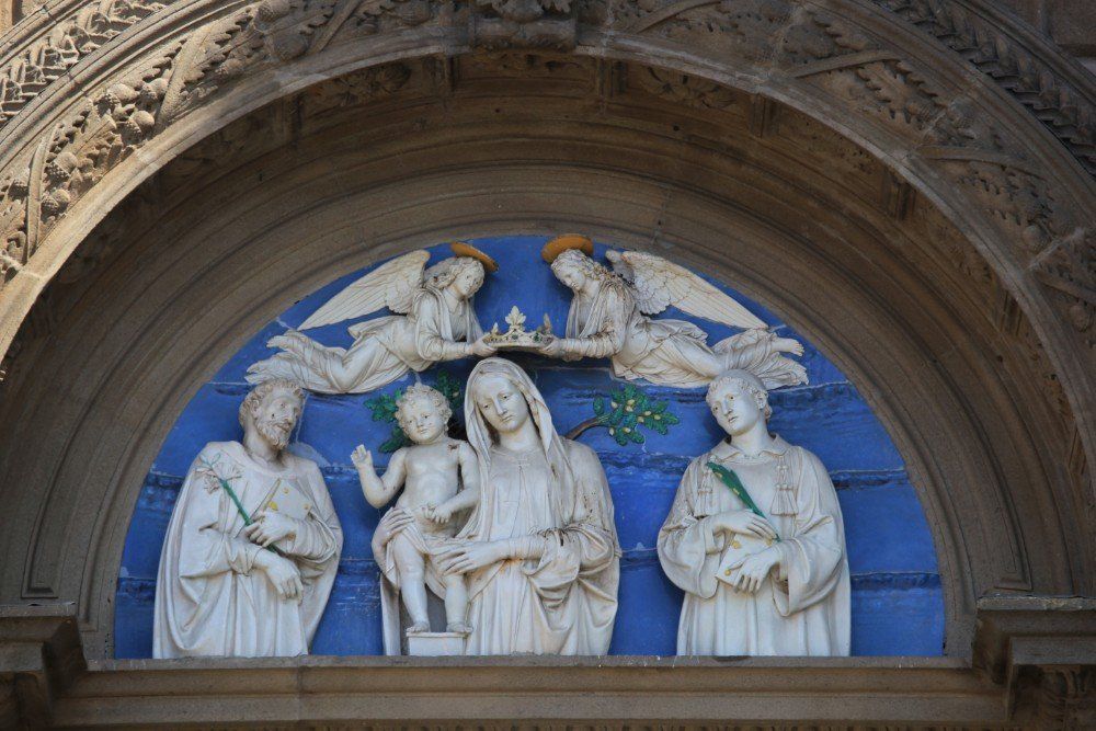 Geformtes Relief der Jungfrau Maria mit Jesus, zwei Engeln, die sie krönen, und zwei Heiligen vor blauem Hintergrund.