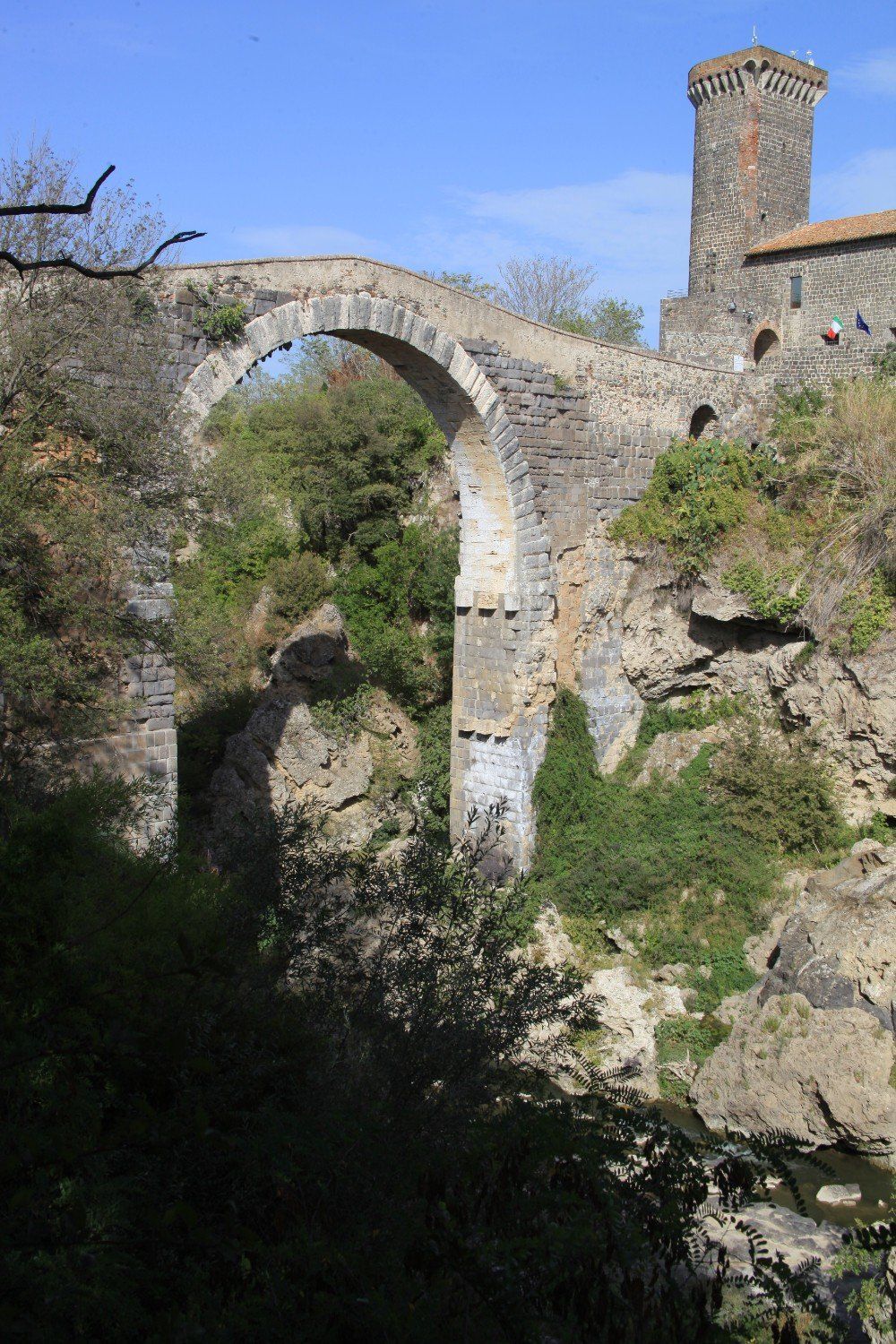 Steinbogenbrücke über eine Schlucht, mit einem Steinturm im Hintergrund und üppigem Grün.