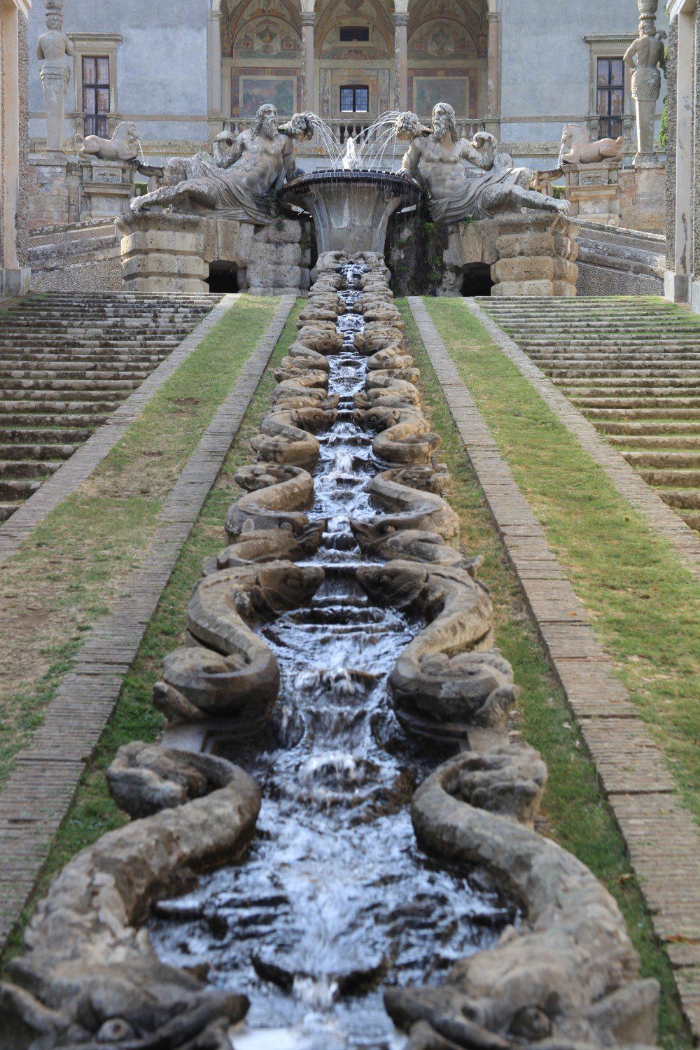 Langer Steinbrunnen mit herabstürzendem Wasser und Skulpturen, der zur kunstvollen Fassade eines Gebäudes führt.