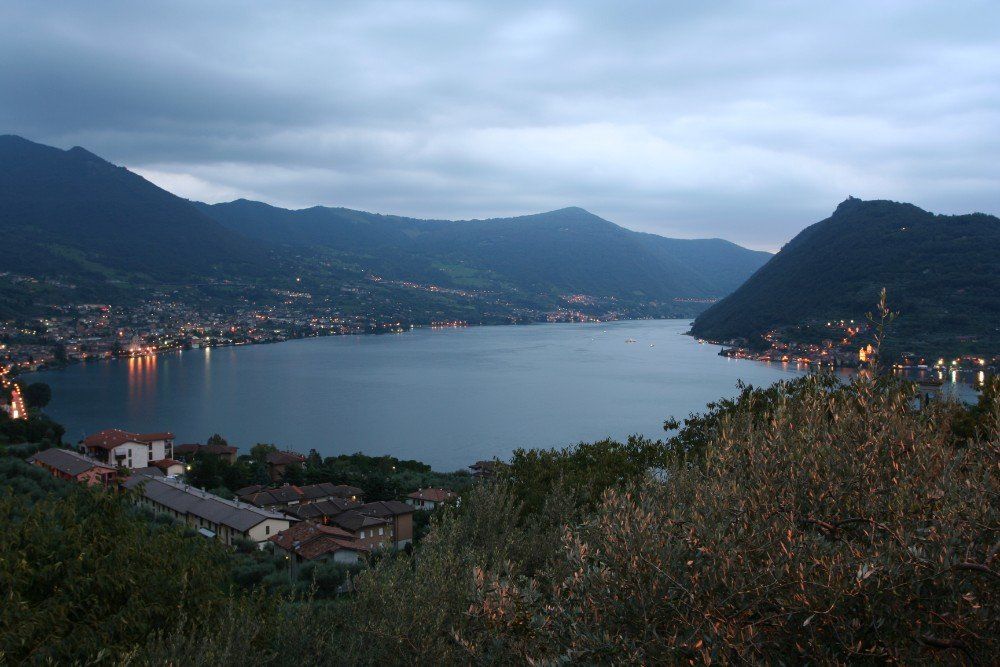 Der Iseosee in der Abenddämmerung, umgeben von Bergen und beleuchteten Städten, unter einem bewölkten Himmel.