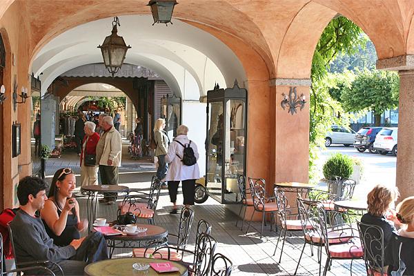 Café mit gewölbtem Gang, in dem Menschen an Tischen sitzen, andere in Geschäften stöbern und Autos entlang der Straße parken.