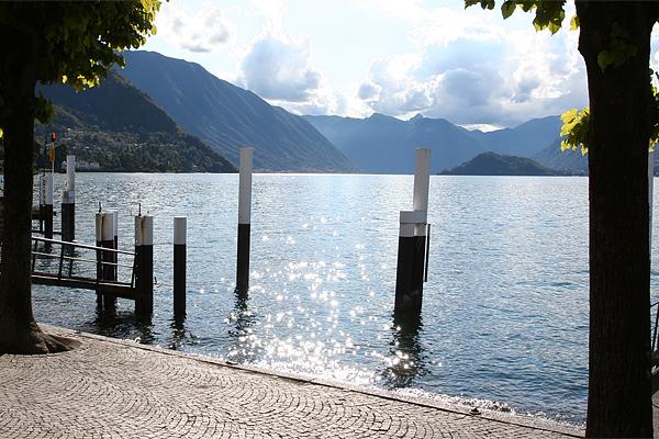 Seeblick mit Holzpfosten im Wasser und Bergen in der Ferne. Die Sonne spiegelt sich im Wasser, mit Bäumen an den Seiten.