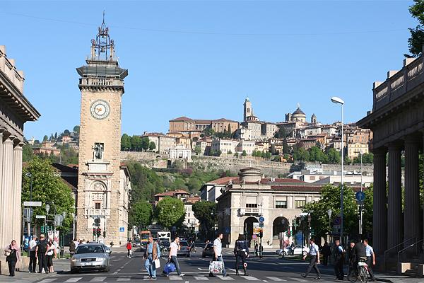 Straßenansicht in Bergamo, Italien, mit einem Uhrturm auf der linken Seite, Menschen, die die Straße überqueren