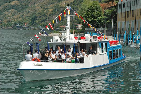 Ein weiß-blaues Ausflugsboot auf einem See, voller Menschen, wehende Fahnen, vor Gebäuden und einem Hügel.