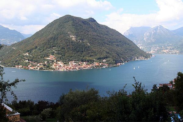 Ein großer, grüner Berg überblickt einen See und ein Dorf mit Gebäuden mit roten Dächern.