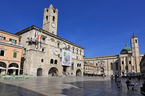 Ein sonniger Tag auf einem großen italienischen Platz mit einem hohen Uhrenturm, historischen Gebäuden und Spaziergängern.