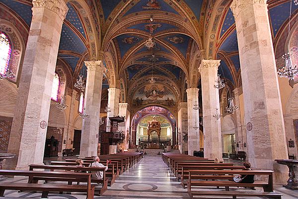 Innenansicht einer Kirche mit hohen Säulen, einer gewölbten Decke mit himmlischen Motiven und Reihen von Holzbänken.