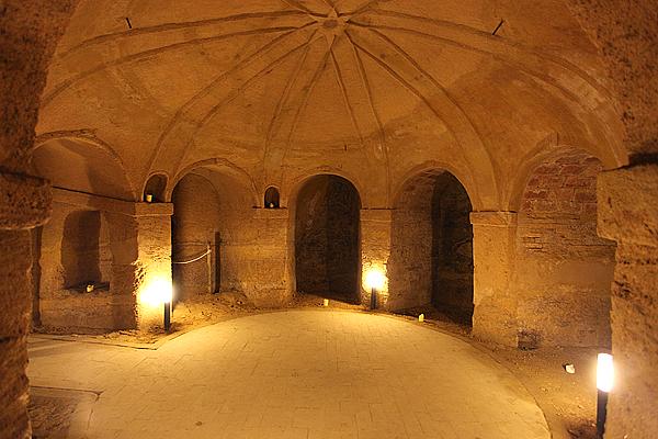 Innenraum einer alten, runden Steinkrypta mit gewölbten Türen und einer gewölbten Decke, beleuchtet von warmen Lampen.