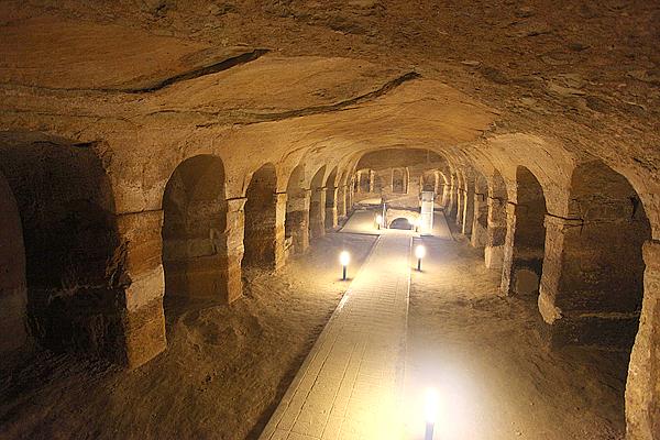 Ein langer, gewölbter unterirdischer Tunnel mit Lichtern säumt den zentralen Weg und ist in den sandfarbenen Fels gehauen.