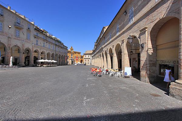 Weitwinkelaufnahme eines gepflasterten Platzes in Italien, gesäumt von gewölbten Gebäuden und Cafés. 