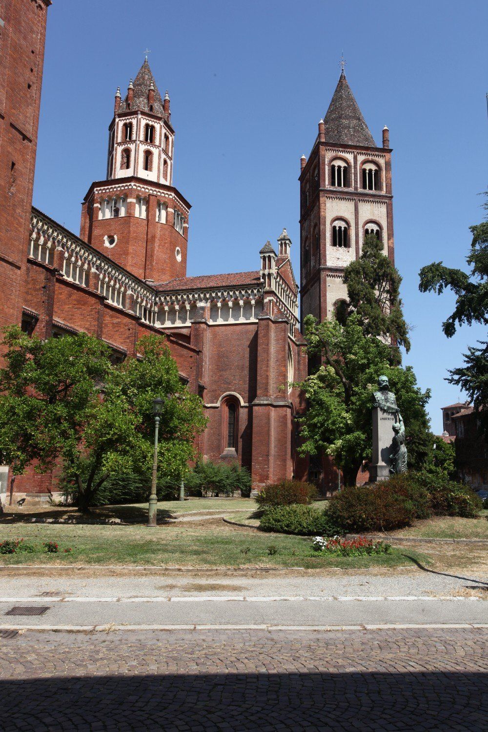 Rote Backsteinkirche mit zwei hohen Türmen vor einem klaren blauen Himmel. Grüne Bäume und Gras im Vordergrund.