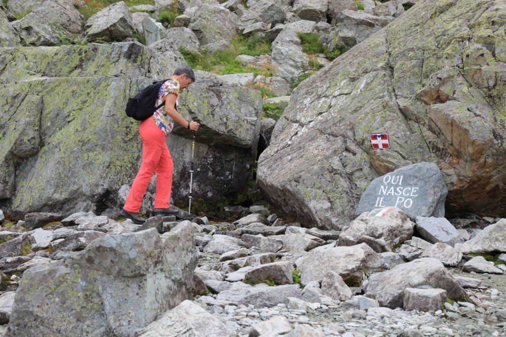Person in roter Hose und Rucksack wandert durch eine felsige Berglandschaft. Wegmarkierung auf einem großen Felsbrocken.