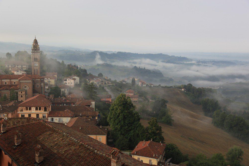 Nebliger Blick auf ein italienisches Dorf mit roten Ziegeldächern, einem hohen Glockenturm und sanften Hügeln in der Ferne.