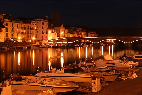 Nachtansicht eines Hafens mit angedockten Booten. Gebäude säumen das Ufer, beleuchtet von im Wasser reflektierten Lichtern. 