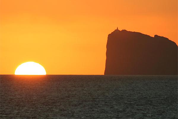 Sonnenuntergang über dem Meer mit einer großen, dunklen Klippe in der Silhouette; orangefarbener Himmel.