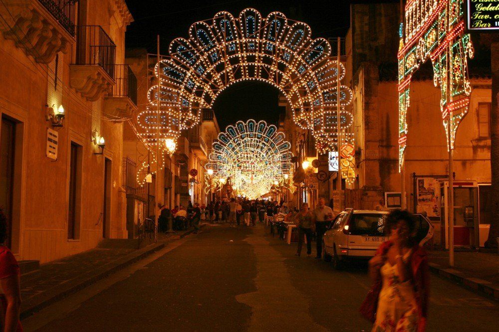 Nachts ist die Straße mit kunstvollen, festlichen Lichterbögen beleuchtet; Passanten und auf der Straße geparkte Autos.