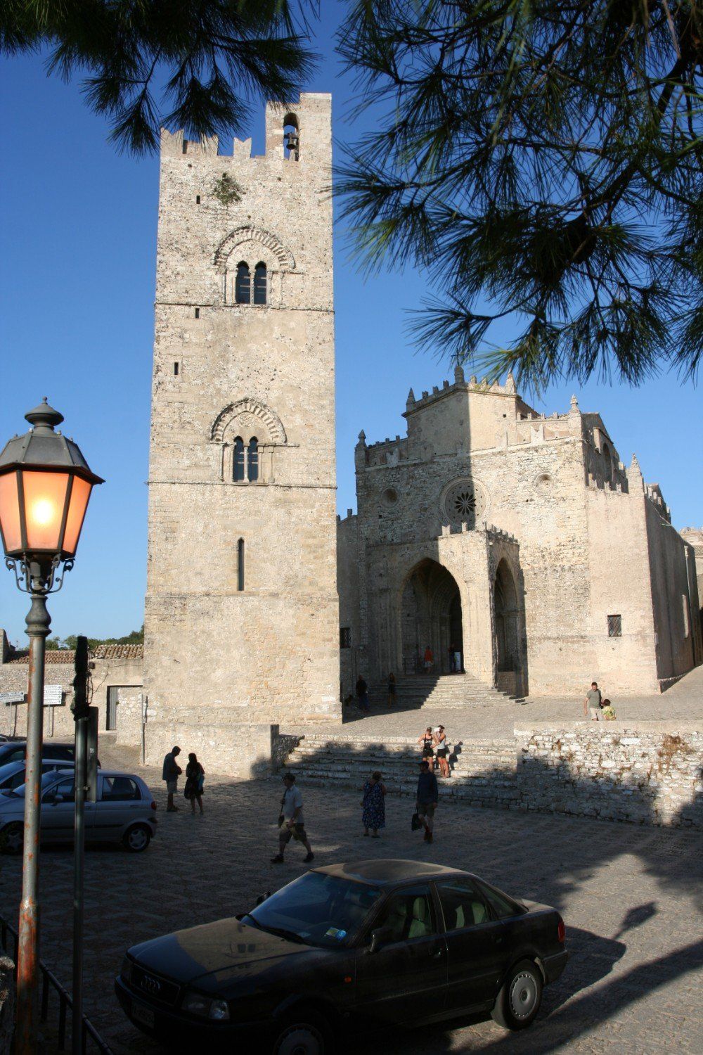 Hoher Steinturm und angrenzendes Gebäude auf einem europäischen Stadtplatz. Sonniger Tag, einige Menschen und Autos.