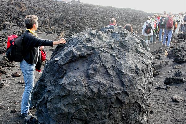 Eine Person zeigt auf einen großen, dunklen Felsen in einer Vulkanlandschaft mit einer Gruppe von Wanderern im Hintergrund.