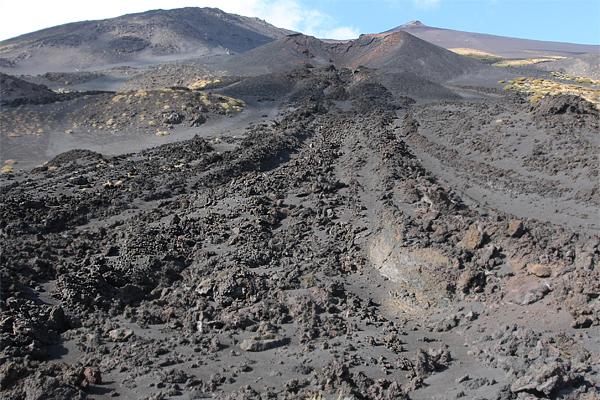Dunkle, felsige Landschaft mit einem Vulkanhang im Hintergrund. Ein Pfad aus dunkler Lava fließt bergab.