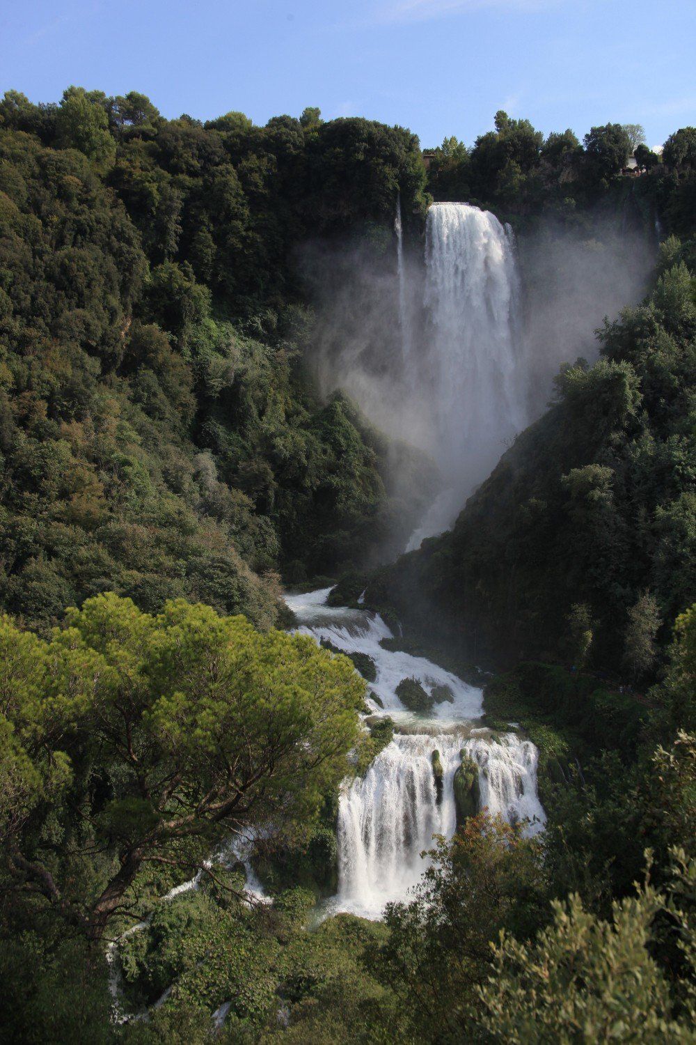 Wasserfall, der einen üppigen, bewaldeten Hang hinabstürzt, mit weißem Wasser vor grünem Laub.