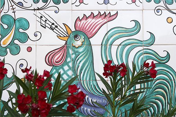 Keramikfliesenkunst eines singenden Hahns in Türkis und Pink, mit Musiknoten, umrahmt von roten Blumen.