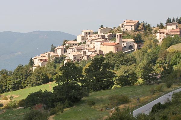 Ein Bergdorf in Italien mit Steinhäusern und Bäumen an einem grünen Hang. Im Vordergrund schlängelt sich eine Straße.