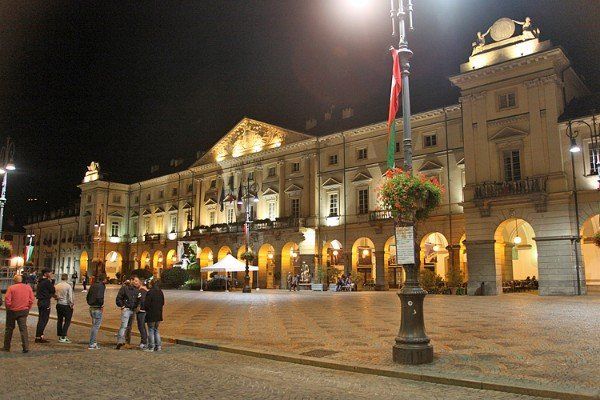 Nachtansicht eines beleuchteten, großen Gebäudes mit Bögen und dekorativen Flaggen auf einem Stadtplatz