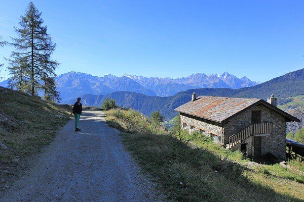 Person, die an einem sonnigen Tag auf einem Schotterweg auf ein Steinhaus zugeht und auf eine Bergkette blickt.