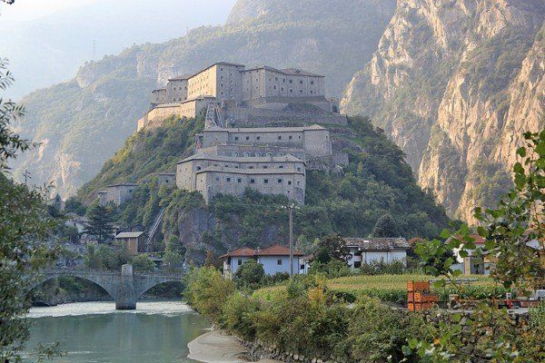 Fort Bard in Italien ist eine mehrstöckige Steinfestung auf einem Berg mit einer Brücke und einem Fluss im Vordergrund.