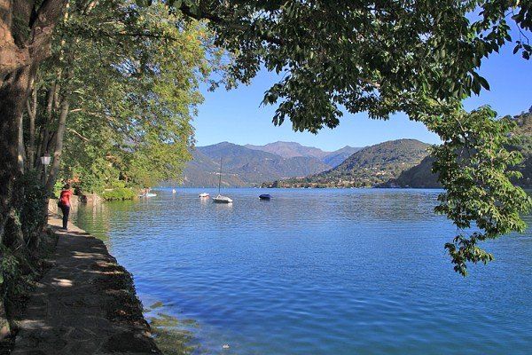 Eine malerische Aussicht auf einen See mit Booten, Bergen und einem Pfad, auf dem eine Person unter Bäumen spazieren geht.