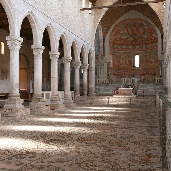 Innenansicht einer alten Kirche mit gewölbten Säulen, Mosaikboden und einer mit Fresken verzierten Apsis.