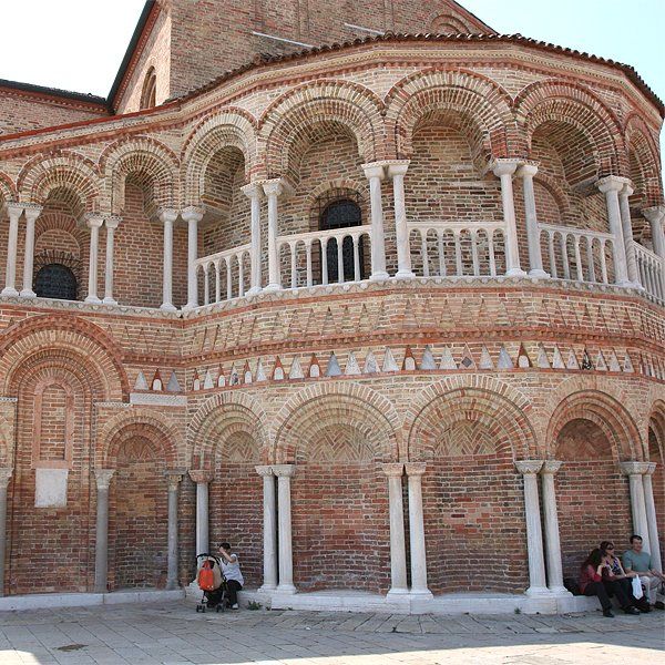 Ein Backsteingebäude mit Bogenfenstern und Säulen in Murano, Italien, vor dem Menschen sitzen.