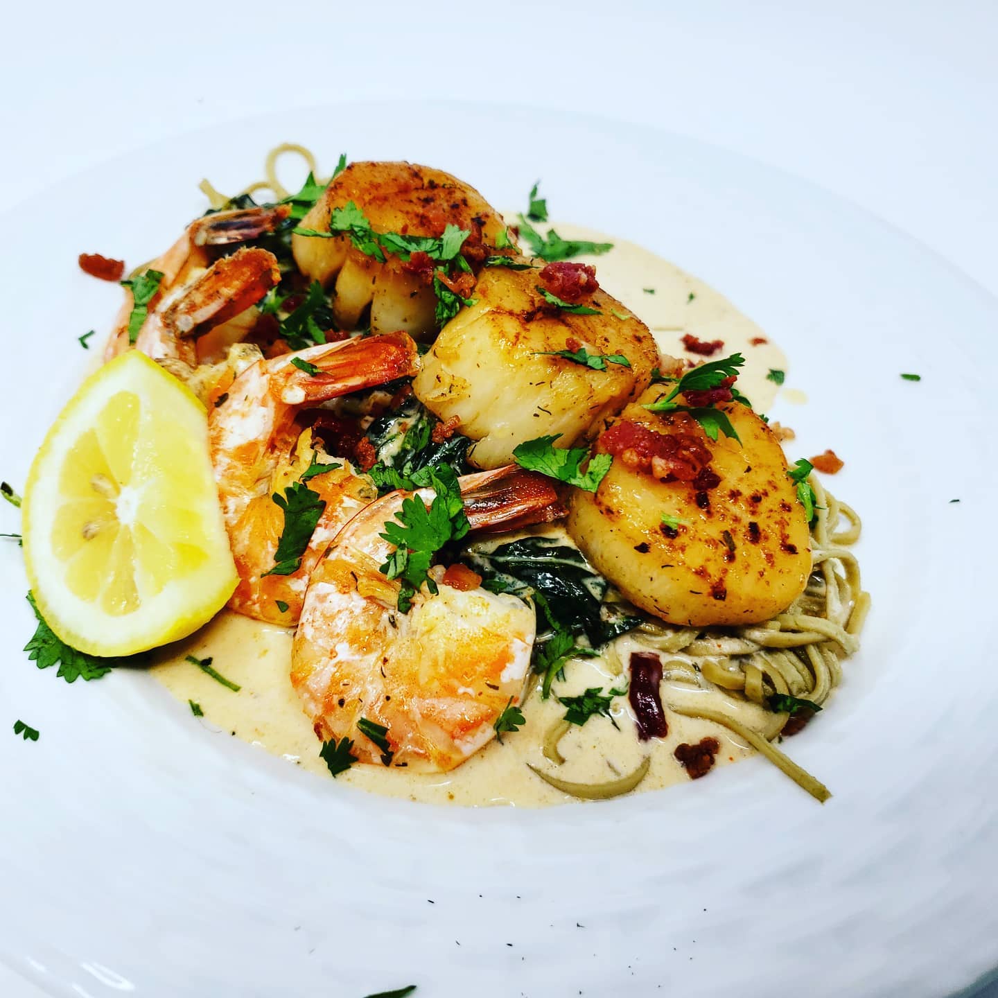 Seared scallops and shrimp on pasta with creamy sauce, garnished with herbs and lemon wedge.