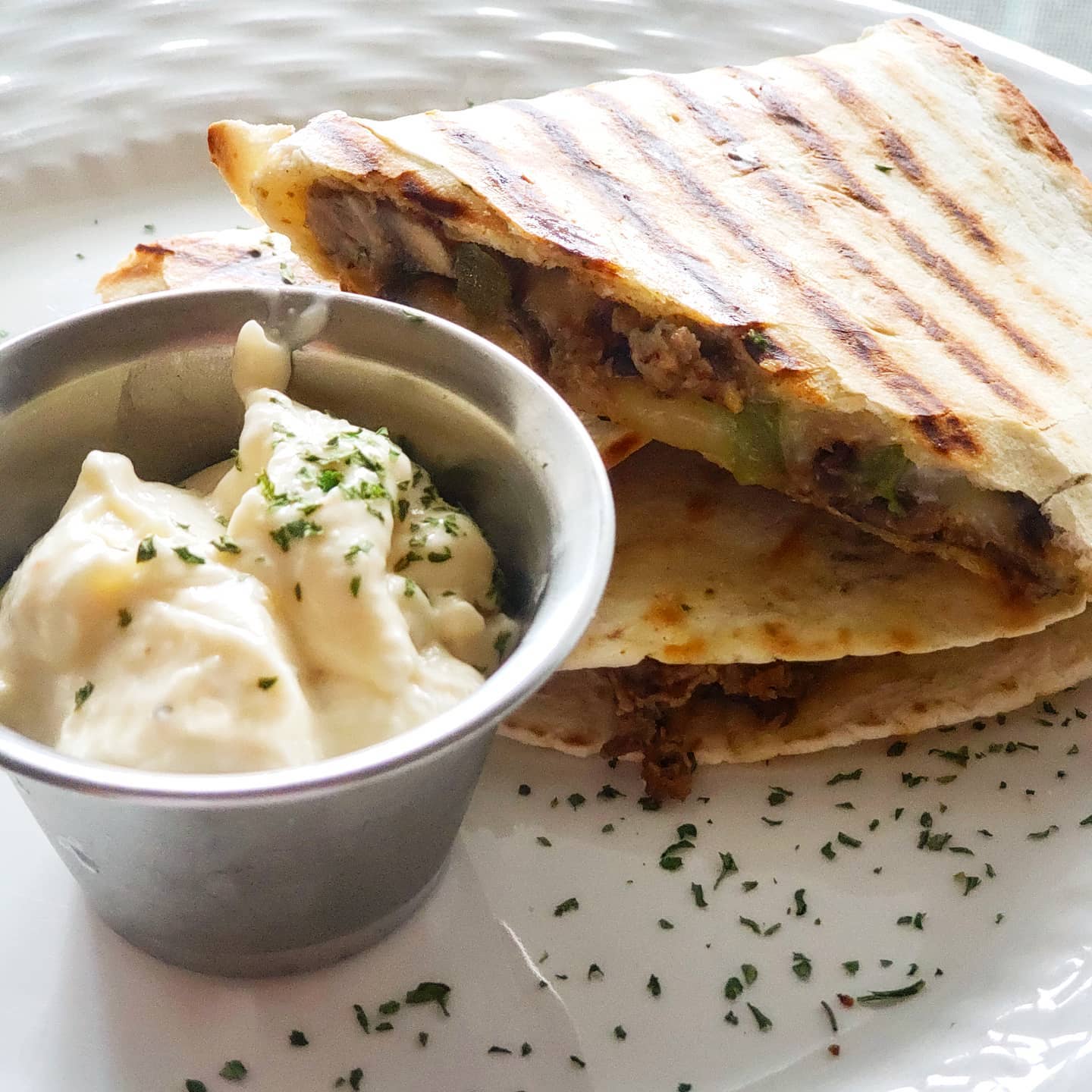 Quesadilla with grilled lines, cut in half, next to a small cup of creamy dip, garnished with parsley on a white plate.