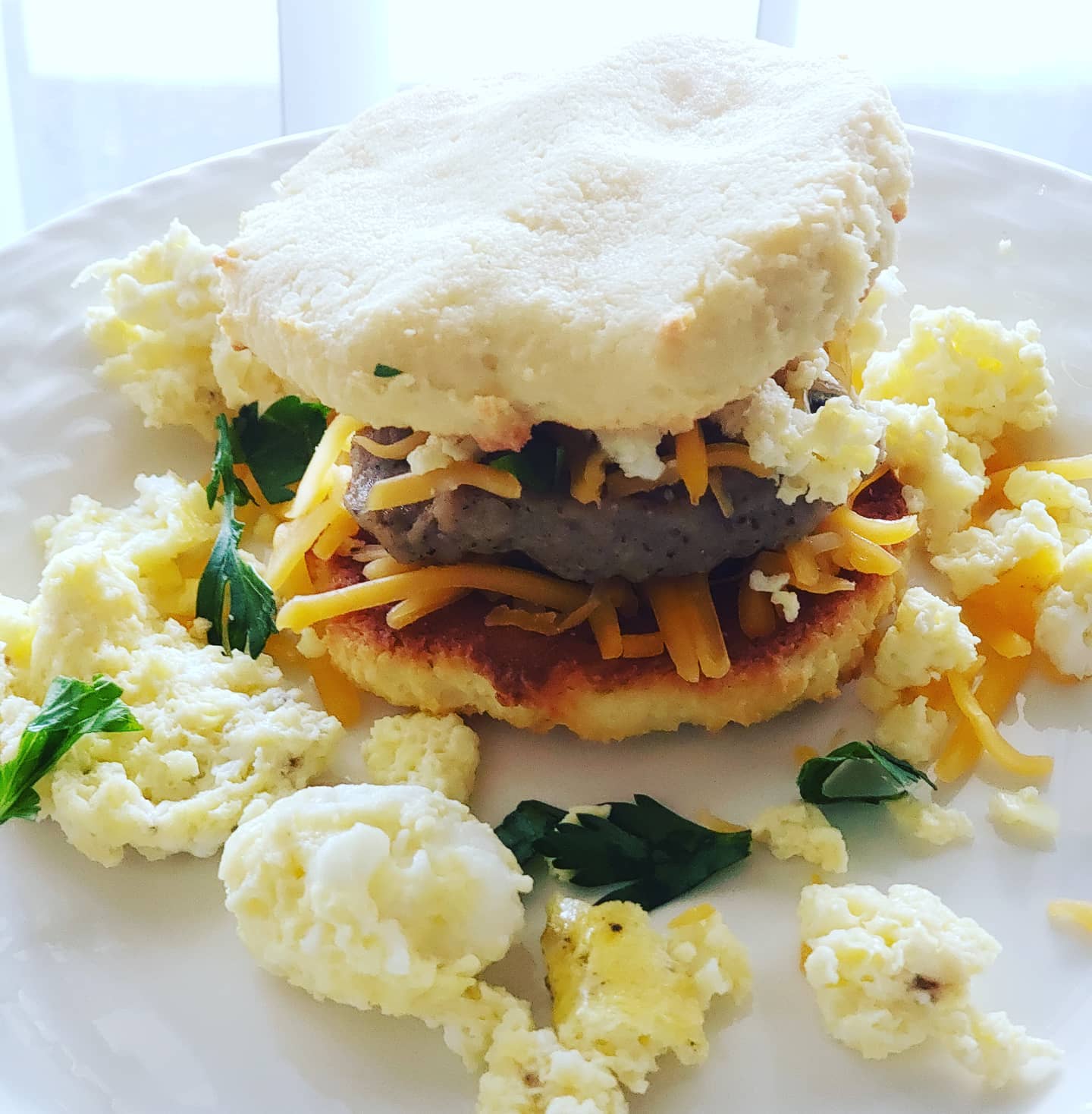 Breakfast sandwich with sausage, cheese, and scrambled eggs between biscuit halves, on a white plate.