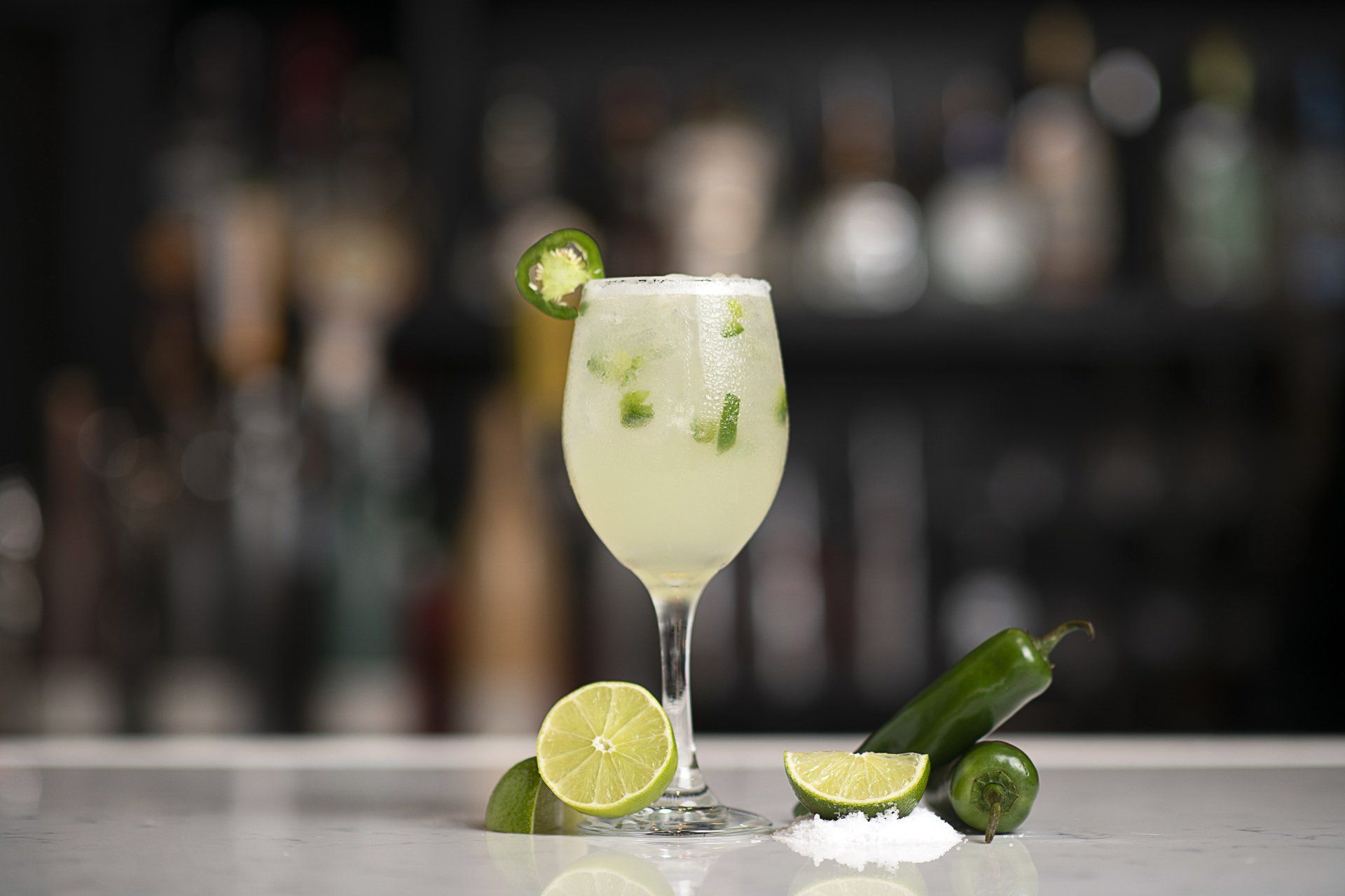 A margarita in a wine glass with limes and jalapenos on a table.