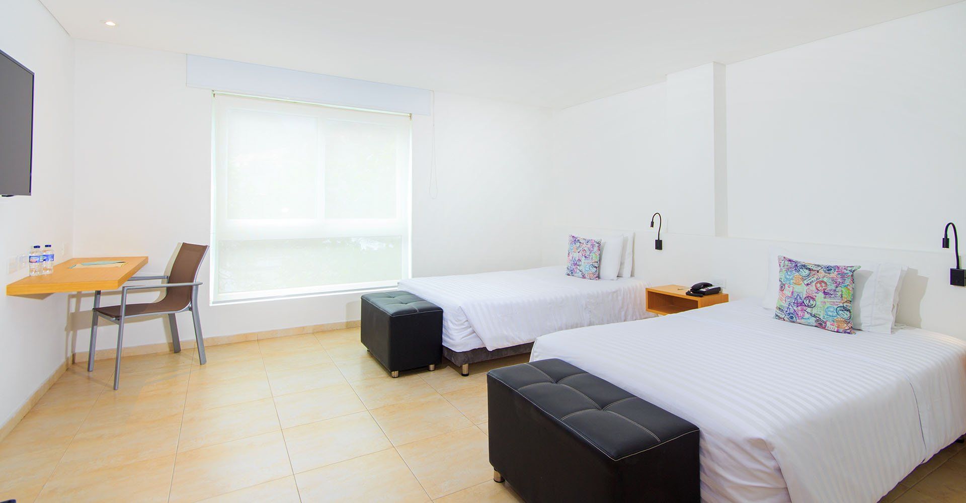 Una habitación de hotel con dos camas individuales, un escritorio y una ventana. La habitación es mayoritariamente blanca, con suelos claros y bancos negros a los pies de las camas.