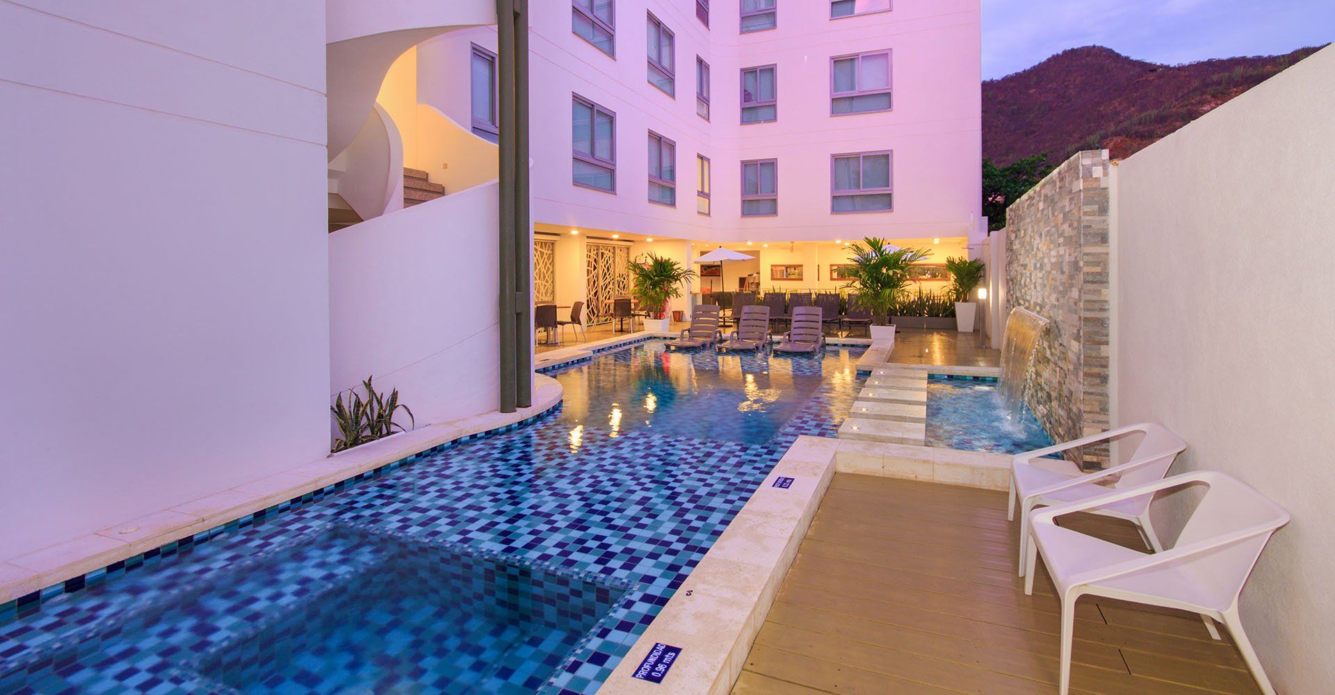 Una piscina de hotel con mosaicos azules, rodeada de paredes blancas y un edificio. Dos sillas blancas dan a la piscina.