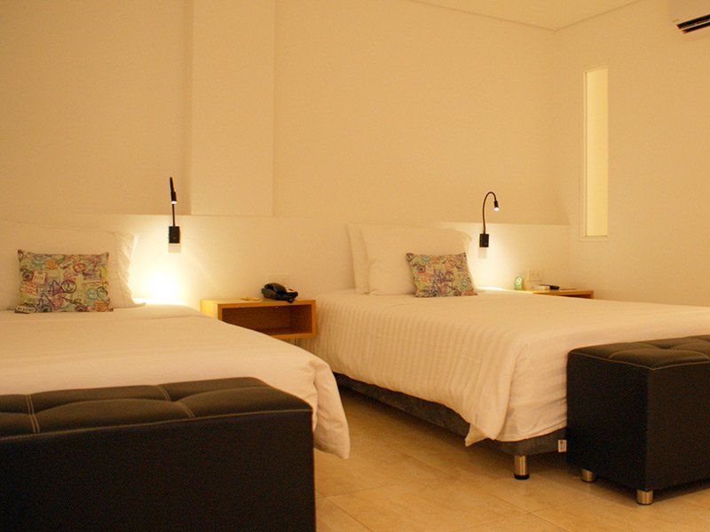 Dos camas individuales en una habitación de hotel con paredes blancas, lámparas de noche y otomanas negras.