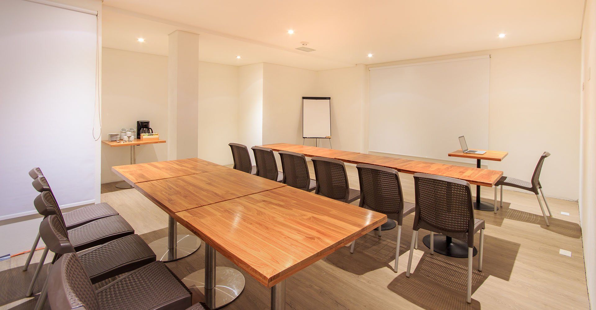 Sala de conferencias con mesas de madera, sillas y pizarra. Cafetera a la izquierda y portátil en una mesita auxiliar.